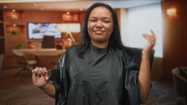 Woman holding scissors with hand and shrugging shoulders in studio set, wearing salon cape and rings, slight smirk and closed eyes; playful confidence.