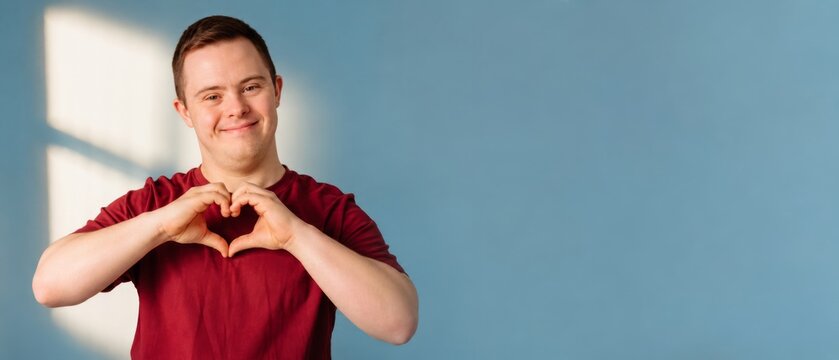 Young man with down syndrome smiling and making heart gesture with hands on a blue banner format