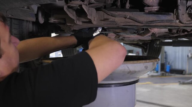 A mechanic working on a car at a repair shop, using a screwdriver to remove a bolt from the underside of the vehicle.