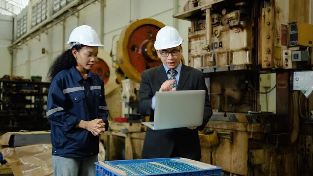 Angry Asian factory manager in a suit and hard hat argues with a female industrial worker over manufactured metal parts on the workshop floor, showing work stress and conflict during a laptop review.