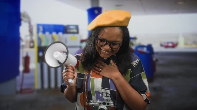 Young woman filmmaker holds megaphone and vintage camera, hand to chest expressing thanks in a street gas station; gratitude creativity.