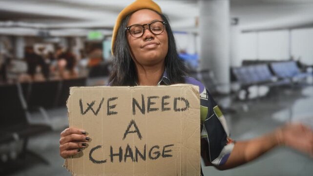 Young african american woman holds cardboard sign 'we need a change' and points finger toward camera in an airport; social change determination.