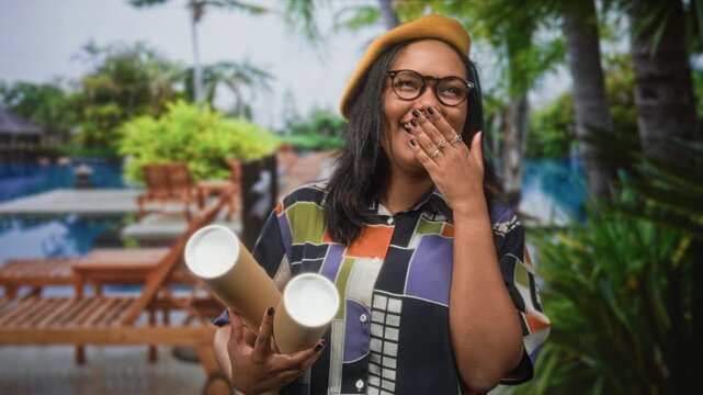 Woman holding rolled blueprints with hand to mouth laughing near pool chairs in building; joy creative.