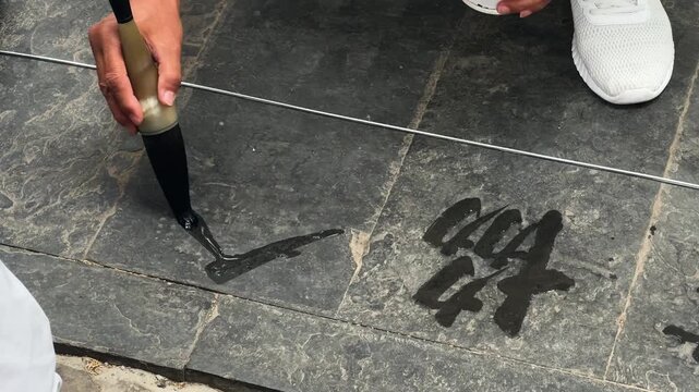 Man calligrapher writing dishu water calligraphy on stone floor with brush. Street artist practicing traditional Chinese ink art. Outdoor performance representing cultural heritage and mindfulness.