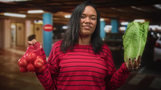 Young woman holds romaine lettuce and mesh bag of tomatoes, hands visible, shrugging gesture in parking building; grocery choice uncertainty.