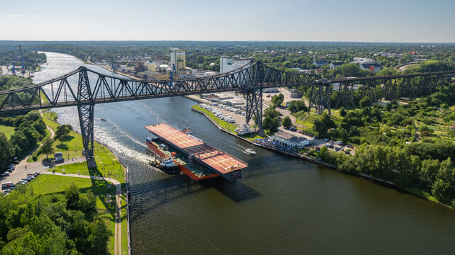 Aerial view of heavy steel bridge deck section on pontoon barge passing under historical Rendsburg High Bridge on Kiel Canal Germany.