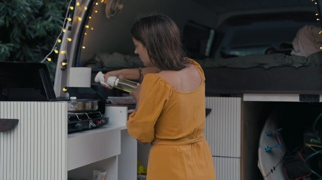 Young adult woman cooks on a stove inside a van, preparing a meal with ingredients on a counter. The van has a cozy interior.