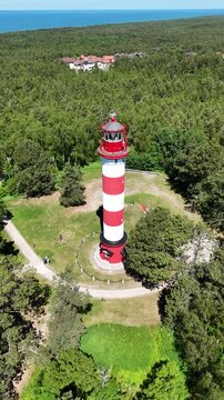 Aerial vertical video of Nida lighthouse on the Curonian Spit, Lithuania. Orbiting drone shot of the red and white striped beacon surrounded by green forest under a clear summer sky.