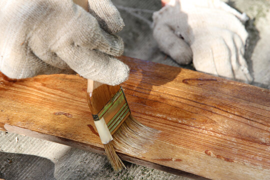 Close-up of a gloved hand applying protective wood stain with a paintbrush to a wooden board outdoors. DIY woodworking, wood preservation, home improvement and renovation concept.