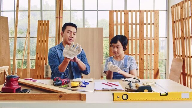 Asian professional carpenter engineer architect woodworker dad amateur young boy sitting wearing safety gloves talking teaching mentoring learning together in housing home building construction site.