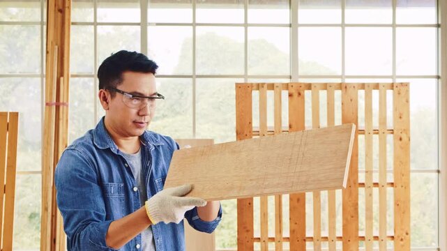 Asian carpentry father sees and checks the wood board in a wood workshop. Wearing safety goggles for woodworking, looking serious and concentrating on work. Carpentry working at a home workshop studio