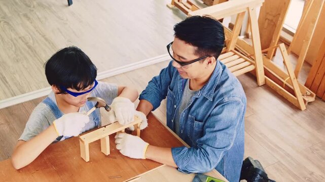Happy Asian father and son work as a woodworker and carpenter. Father teaching his son to hammer nails on a wooden plank carefully together. carpentry working at a home workshop studio.