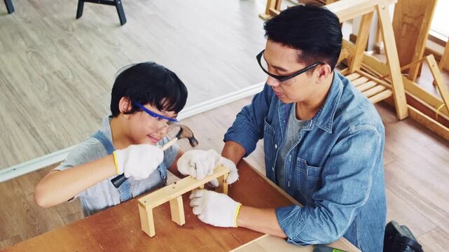 Happy Asian father and son work as a woodworker and carpenter. Father teaching his son to hammer nails on a wooden plank carefully together. carpentry working at a home workshop studio.