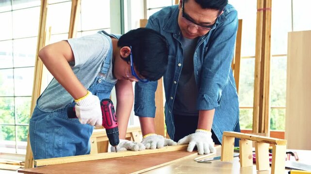 Asian father and son work as a woodworker or carpenters. Close up hands of the father and his son drill holes in a wooden plank carefully together. carpentry working at a home workshop studio.