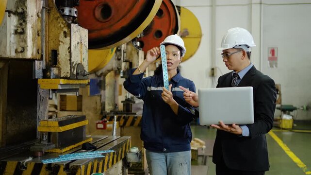 Asian female factory worker and male manager engineer in hard hats inspect a metal part near a stamping press machine. Quality control discussion with laptop in an industrial manufacturing plant.