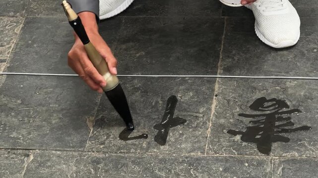 Person calligrapher practicing water calligraphy on stone pavement with large brush. Street artist writing Chinese characters with water. Traditional oriental art, mindful hobby and zen practice.