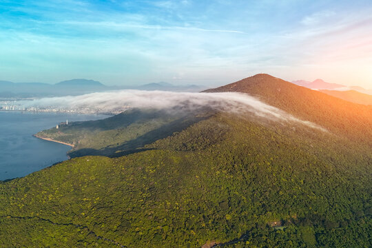 Clouds float over the dense jungle and rugged mountains of Son Tra Peninsula in Danang as morning sunlight hits the coastal landscape near the calm blue ocean