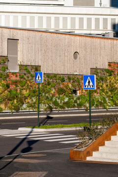 A pedestrian zebra crossing links the urban street and road, while blue signs stand beside concrete stairs beneath a textured facade designed for safety
