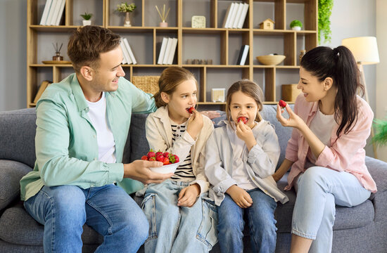 Family eating strawberries together at home. Smiling parents and children share a healthy snack on the sofa, enjoying fresh fruit in a bright living room. Healthy family lifestyle and bonding.