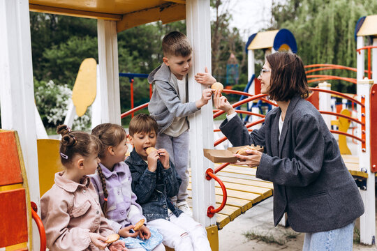 Smiling happy woman teacher sharing cookies with children at playground