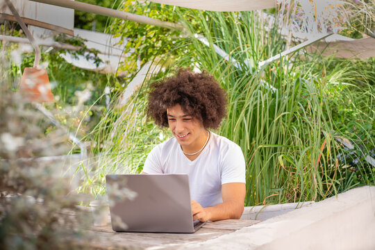 Smiling young ethnic men , dressed in casual clothes, works on a laptop outdoors, in a green area in nature. Green open space for coworking, freelancer, relaxing, coffee, food.Free place to work