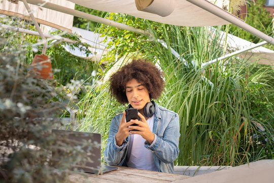 Young mixed race boy ,dressed in casual clothes using smartphone in green area in nature. Green open space for coworking, freelance, relax, coffee, food. Free place to work, business, study, freelance