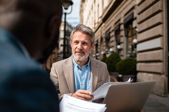 Senior businessman meeting client with laptop and documents at outdoor city cafe