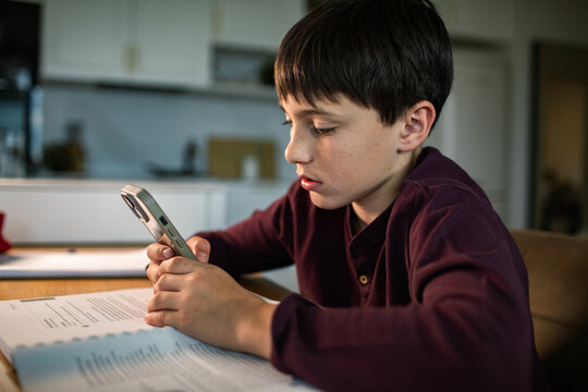 Preteen boy using smartphone while doing homework at kitchen table