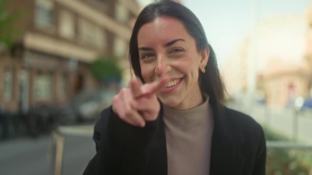Young hispanic woman points finger at camera on street while smiling and flashing peace sign in blazer; playful invitation.