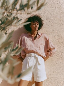 Woman in dusty rose satin oversized shirt and white shorts poses against a textured peach wall, with olive branches framing the scene in a bright outdoor setting