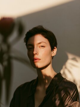 Close up portrait of a woman with short dark hair illuminated by soft sunlight, casting shadows on her face against a light background with plant silhouettes