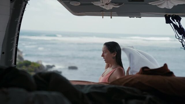 A woman stands by the ocean, wrapped in a towel after swimming, next to her camper van with waves in the background.