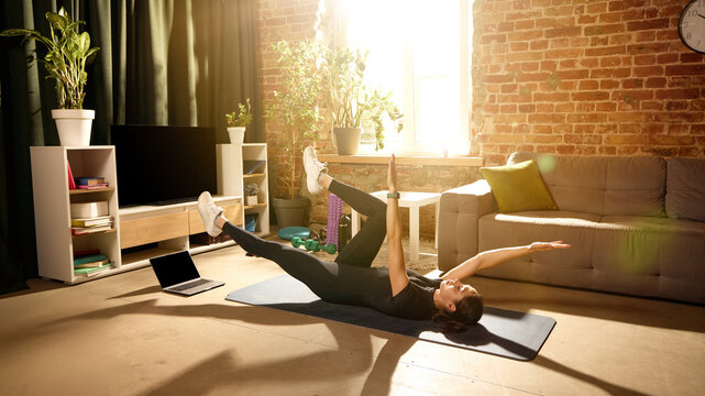 Woman doing dead bug core exercise on mat beside laptop at home. Concept of online fitness classes, abdominal strength, posture support, coordination training and effective home workout routines.