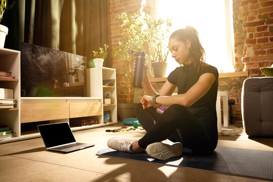 Woman checking smartwatch and holding water bottle after home workout. Concept of fitness tracking, hydration habits, workout recovery, wearable technology and monitoring personal health progress.