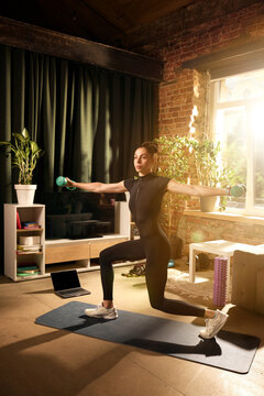 Woman performing weighted lateral raise in kneeling lunge position during home strength workout. Concept of shoulder strength, functional fitness, home exercise, muscle training and healthy lifestyle.