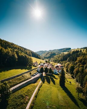 Sunny village in green valley with forested hills for travel and rural tourism, clear blue sky
