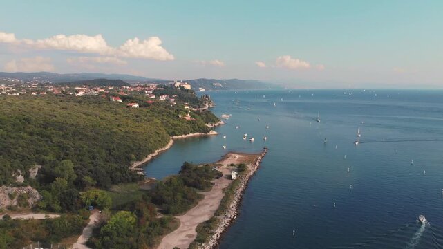 Aerial 2.7K 60fps drone shot of the scenic Adriatic coast near Villaggio del Pescatore, Italy, with Duino Castle on the cliffs and sailboats on the blue sea