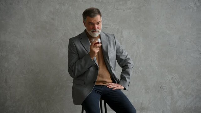 Handsome and successful businessman in stylish suit holding glass whiskey while sitting on the stool