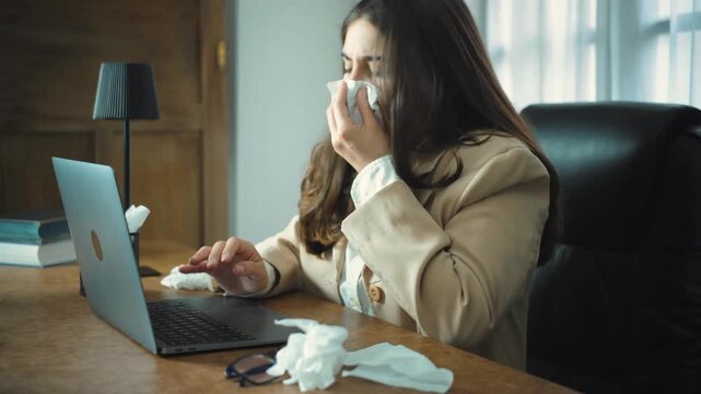 Young Hispanic businesswoman with a fever struggling to finish her work on a laptop in a modern office, showing exhaustion, illness, and dedication during the workday.
