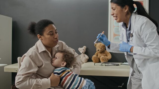 Young Hispanic female pediatrician using teddy bear to comfort toddler on Biracial mothers lap during appointment in clinic. Video is perfect for promoting pediatric and family healthcare