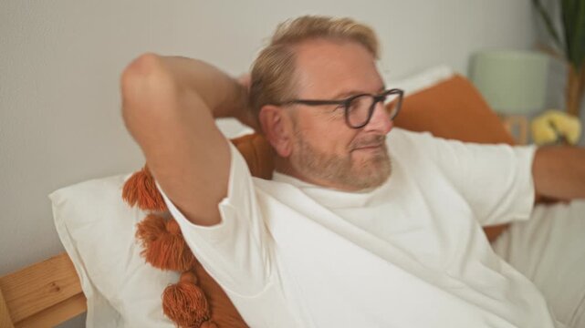 Man with glasses reclining on bed with hands behind head, orange pillows and wooden frame visible in a building; contentment rest.