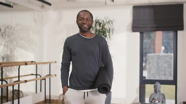 African American yoga man posing at camera. Male yoga coach holding fitness mat. Professional yoga instructor standing at studion and smiling at camera. Cheerful yoga trainer satisfied with session.