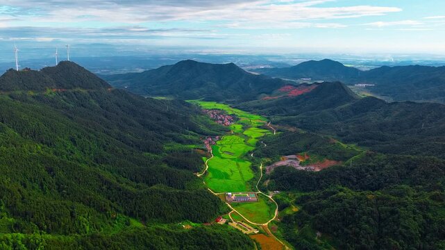 Aerial view of lush green mountain valley with terraced agricultural fields, winding road, small village and wind turbines on the ridge under a blue sky.
