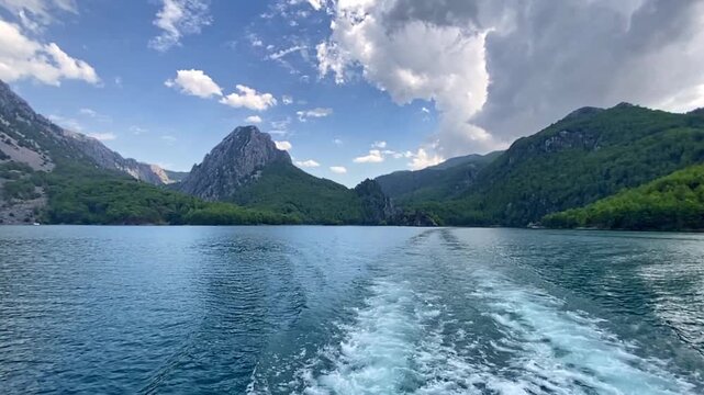 Scenic view of a tranquil lake reflecting the surrounding mountains and lush greenery under a partly cloudy sky, with visible boat wake creating ripples on the water surface