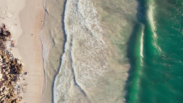 Drone view of waves roll gently onto a sandy beach as sunlight reflects on the clear water during midday.