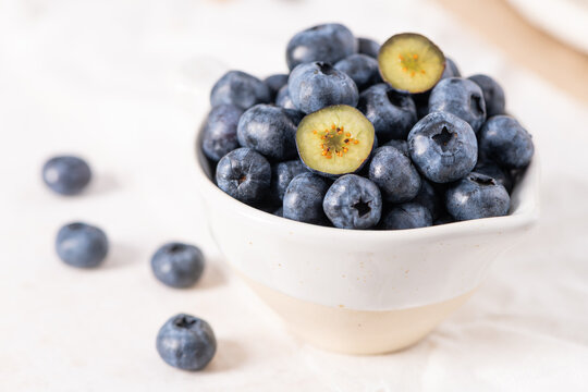 Fresh blueberries are piled into a small rustic ceramic bowl, with a few sliced berries revealing their green centers on a white fabric surface