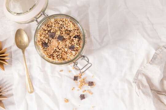 Crunchy granola with dark chocolate chunks in a glass jar on a white cloth for a healthy and nutritious morning breakfast meal
