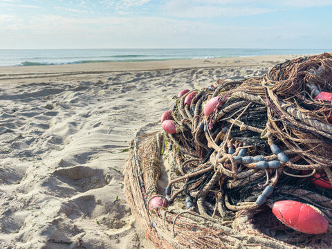 Traditional fishing nets with red floats resting on a sandy beach in Portugal with the calm ocean waves and blue sky in the background under bright morning light
