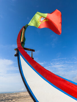 A close up of a traditional Arte Xavega boat bow with the Portuguese flag waving against a bright blue sky at a coastal beach in Ovar Portugal