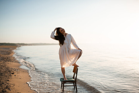 Young woman standing on chair in sea water alone wearing white linen dress over sun light and seashore. Loneliness.